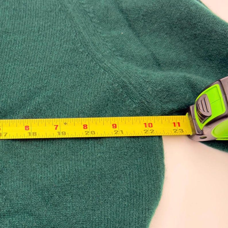 Puede incluir: Un su&eacute;ter de punto verde oscuro con una costura visible a lo largo del hombro. Una cinta m&eacute;trica amarilla est&aacute; colocada sobre la tela, indicando una medida de ancho que se extiende de aproximadamente 13 cm a 28 cm en la escala m&eacute;trica.