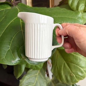 May include: A white ceramic mug with a black handle and the text "But First, Coffee" in a bold, black font. The mug is set against a neutral background, highlighting its simple design.
