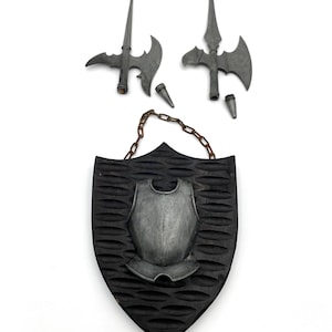 May include: A medieval-themed decorative set featuring a black shield with a carved texture and a metallic breastplate. Two gray polearms and two conical pieces are positioned above the shield, and a chain hangs from the shield's top.