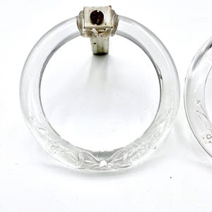 May include: Clear glass drawer pulls with a floral design etched into the glass. The pulls have a circular shape with a metal mounting bracket. The hardware is attached to the glass with a screw.