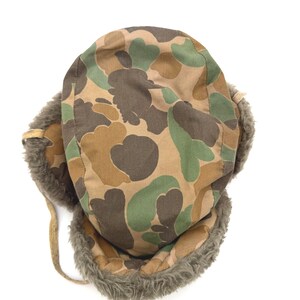 May include: A trapper hat featuring a camouflage pattern in shades of brown, tan, and green. The hat is lined with thick, textured gray faux fur along the ear flaps and front brim, and includes thin tan fabric ties attached to the side flaps.