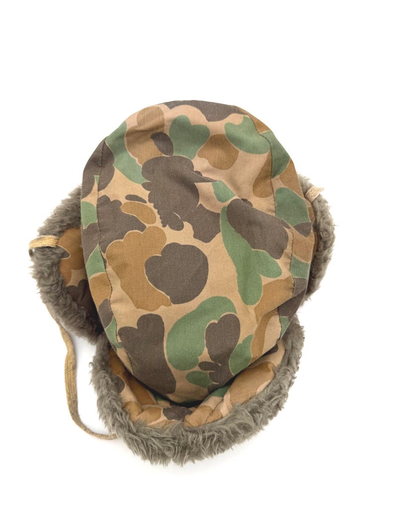 May include: A trapper hat featuring a camouflage pattern in shades of brown, tan, and green. The hat is lined with thick, textured gray faux fur along the ear flaps and front brim, and includes thin tan fabric ties attached to the side flaps.