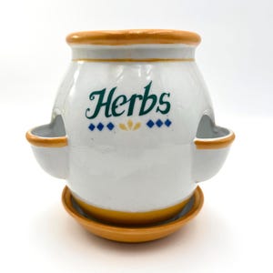 May include: White ceramic herb planter with a yellow rim and base. The planter has two side openings and the word "Herbs" in green script. Decorative blue and yellow accents are below the word.
