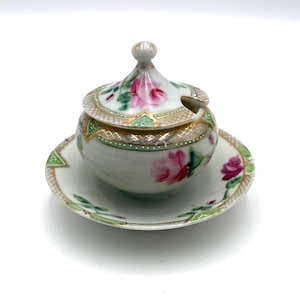 May include: A small, antique porcelain sugar bowl with a lid and saucer. The bowl and lid are decorated with pink roses, green leaves, and gold trim. The saucer has a matching floral design.