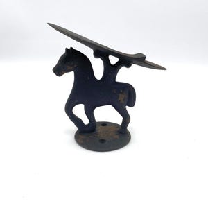 May include: A dark, weathered cast iron boot jack shaped like a horse standing on a circular base. The horse figure supports a long, narrow, angled metal plate used for removing footwear, featuring visible signs of rust and aged metal texture.