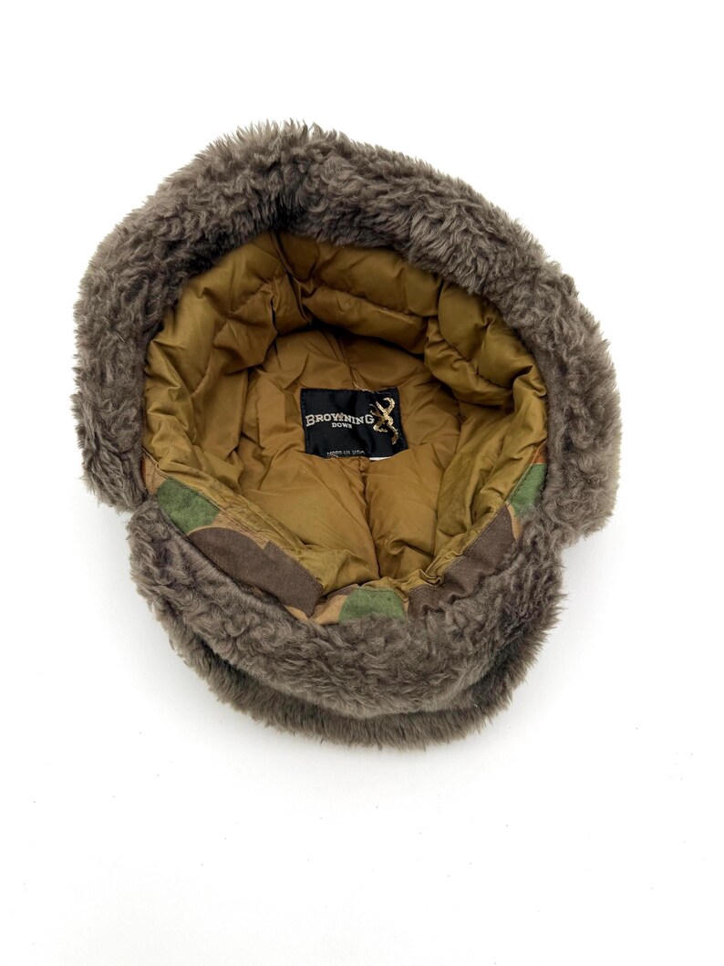 May include: A brown trapper hat with a thick, fuzzy faux fur lining and a quilted tan interior. The hat features a camouflage pattern on the outer band and a black label inside with the text Browning Down and a buck logo, stating Made in USA.