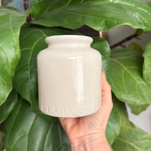 May include: A small, cylindrical, off-white ceramic jar with a smooth, glossy finish and a short, narrow neck. The base of the jar features the embossed text "LAB Lagre" in capital letters, providing a subtle, tactile detail to the design.