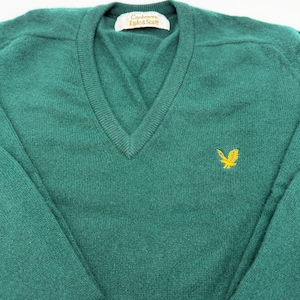 Puede incluir: Un su&eacute;ter de color verde oscuro con cuello en V, mangas largas y cuello acanalado. La prenda presenta un logotipo de &aacute;guila bordado en amarillo en el pecho izquierdo y una etiqueta interior blanca con el texto Cashmere Lyle & Scott en negro.