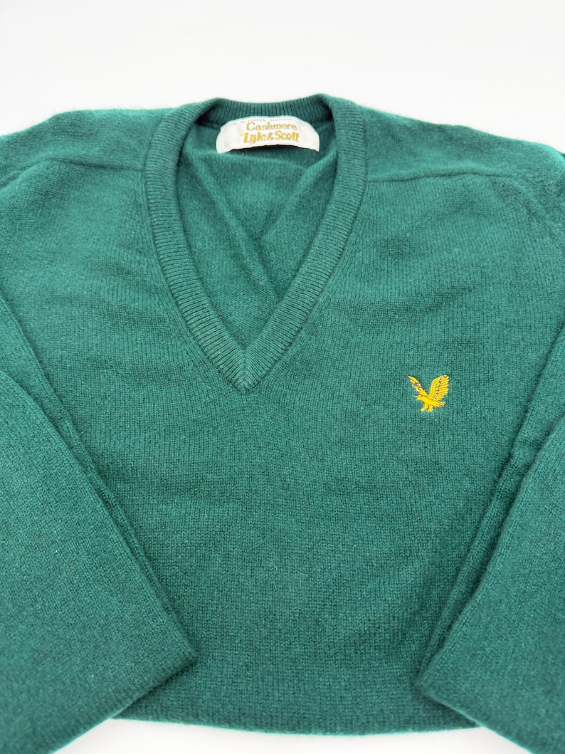 Puede incluir: Un su&eacute;ter de color verde oscuro con cuello en V, mangas largas y cuello acanalado. La prenda presenta un logotipo de &aacute;guila bordado en amarillo en el pecho izquierdo y una etiqueta interior blanca con el texto Cashmere Lyle & Scott en negro.