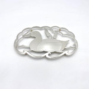 May include: A silver-colored duck-shaped decorative item. The duck is depicted swimming in water, surrounded by a cloud-like frame. Reeds are visible behind the duck. The item is likely a brooch, pin, or decorative element.
