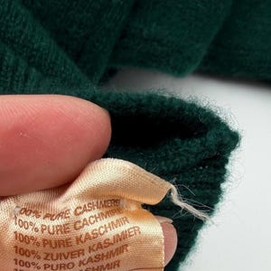 Puede incluir: Una prenda de punto de color verde oscuro con una etiqueta de tela clara que dice 100% PURE CASHMERE, 100% PURE CACHMIRE, 100% PURE KASCHMIR, 100% ZUIVER KASJMIER, 100% PURO KASHMIR, 100% REN KASHMIR e indica que est&aacute; fabricada en Escocia.