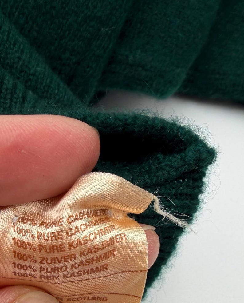 Puede incluir: Una prenda de punto de color verde oscuro con una etiqueta de tela clara que dice 100% PURE CASHMERE, 100% PURE CACHMIRE, 100% PURE KASCHMIR, 100% ZUIVER KASJMIER, 100% PURO KASHMIR, 100% REN KASHMIR e indica que est&aacute; fabricada en Escocia.