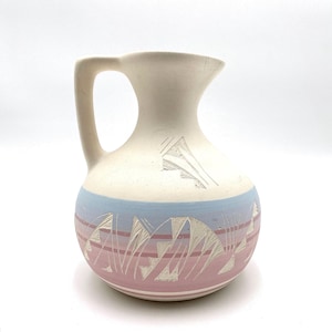 May include: A cream-colored ceramic pitcher with a handle and spout. The pitcher features a blue and pink band with etched geometric designs. The top portion has etched mountain-like shapes.