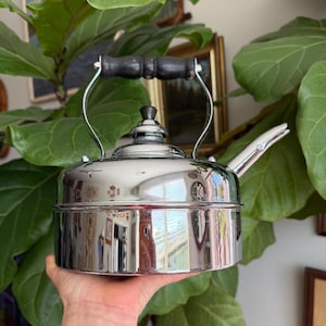 May include: A shiny, chrome-plated tea kettle with a black wooden handle and spout. The kettle has a classic design, with a rounded body and a lid with a small knob. The handle is arched and attached to the kettle with metal supports.