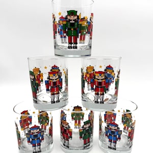 May include: Six clear drinking glasses with a festive nutcracker design. Each glass features a colorful illustration of nutcracker soldiers in red, blue, and green uniforms. The glasses are arranged in a pyramid shape, perfect for holiday gatherings.