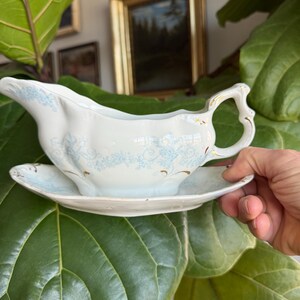 May include: A white porcelain gravy boat with a matching saucer. The boat features a delicate blue floral pattern and gold trim. The handle is ornate. The set is displayed against a backdrop of green foliage and framed artwork.