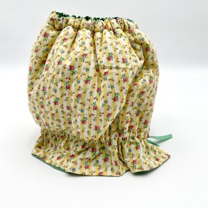 May include: A yellow and white fabric with a floral pattern and green trim. The fabric is gathered at the top and has a green ribbon tied at the bottom.