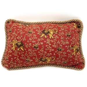 May include: Rectangular red pillow with a repeating pattern of gold elephants and floral designs. The pillow has a rope trim and is likely used for decorative purposes. The elephants are detailed with brown accents.