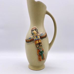 Puede incluir: Una jarra de cerámica verde claro con una superficie texturizada. La jarra tiene un asa y un diseño de tótem nativo americano con un pájaro en la parte superior.