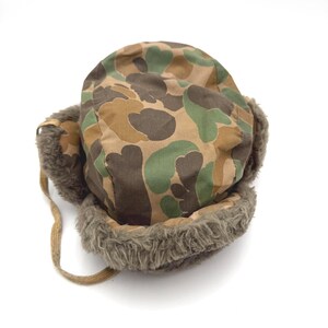 May include: A trapper hat featuring a camouflage pattern in shades of brown, tan, and green. The hat includes ear flaps lined with thick, textured brown faux fur and a matching brown fabric chin strap hanging from the side of the headpiece.