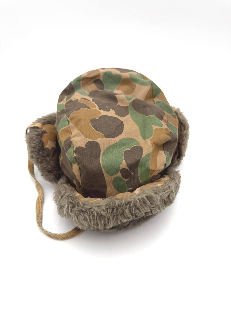 May include: A trapper hat featuring a camouflage pattern in shades of brown, tan, and green. The hat includes ear flaps lined with thick, textured brown faux fur and a matching brown fabric chin strap hanging from the side of the headpiece.