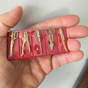 May include: A set of five miniature gold-toned metal tool charms arranged in a red plastic display tray. The collection includes a pair of scissors, pliers, a screwdriver, tin snips, and a folding pocket knife, each secured in individual slots.