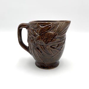 May include: A brown ceramic pitcher with a handle and a detailed relief design of leaves and a pinecone. The pitcher has a slightly flared rim and a pedestal base. The dark brown color gives it a vintage or rustic feel.