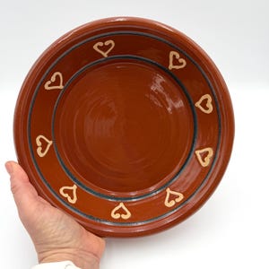 May include: A terracotta-colored ceramic plate with a heart design. The plate has a wide rim with a dark blue and tan heart pattern. The plate is held by a person's hand against a white background.
