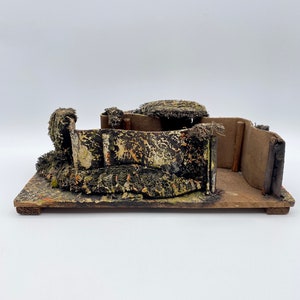 May include: A miniature model of a bunker made of cardboard and painted with camouflage colors. The bunker has a small opening and is surrounded by green and brown foliage.