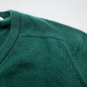 Puede incluir: Un su&eacute;ter de color verde esmeralda oscuro con cuello redondo acanalado y una costura visible en el hombro. El tejido de punto tiene una textura suave y ligeramente difusa en toda la prenda, destacando el color verde intenso y las costuras.