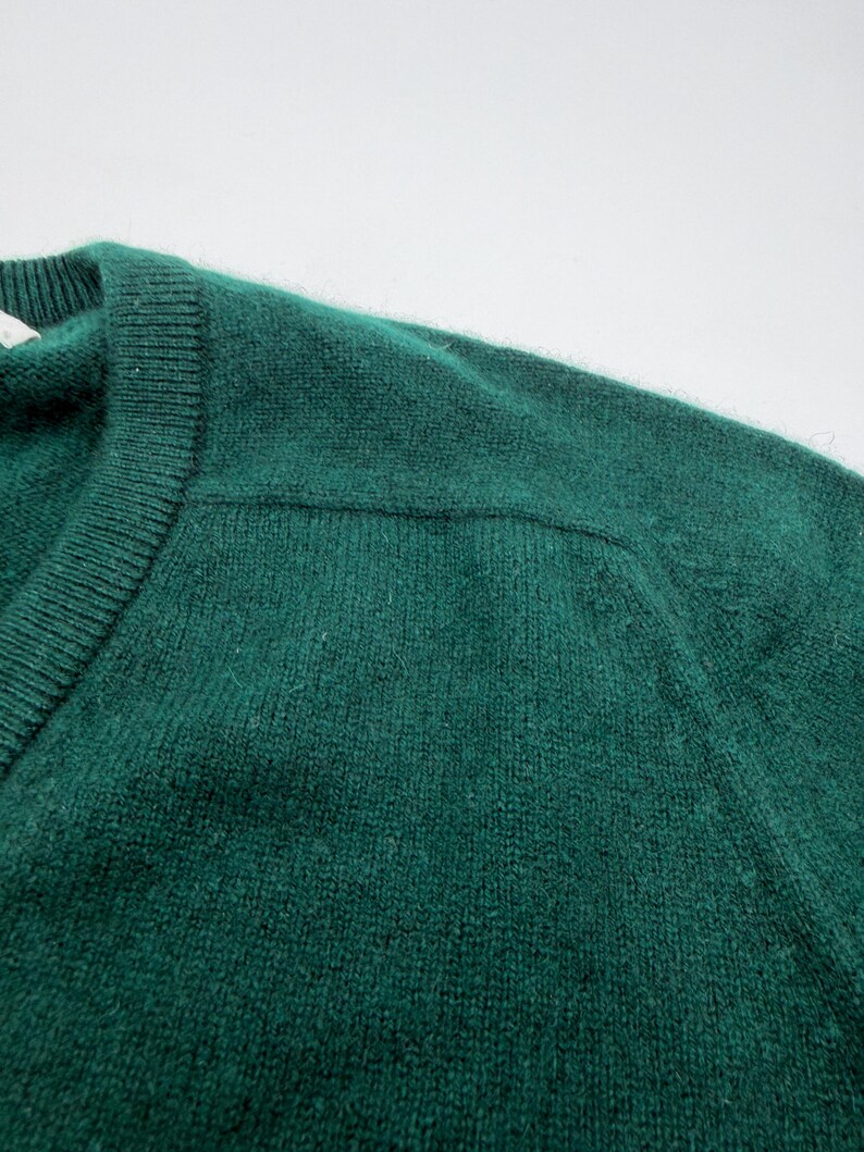 Puede incluir: Un su&eacute;ter de color verde esmeralda oscuro con cuello redondo acanalado y una costura visible en el hombro. El tejido de punto tiene una textura suave y ligeramente difusa en toda la prenda, destacando el color verde intenso y las costuras.