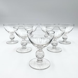 May include: Set of clear, vintage-style stemmed glasses. Each glass features a round bowl, a decorative ball-shaped stem, and a circular base. The glasses are arranged on a white surface, showcasing their elegant design.