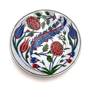 May include: A round, white ceramic plate with a silver rim. The plate features a floral design with red, blue, and green accents. The flowers and leaves are intricately detailed, creating a decorative piece.