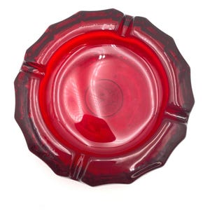 May include: A vintage, ruby-red glass ashtray with a scalloped edge. The ashtray has a circular design with four grooves for resting cigarettes. The glass is transparent, allowing light to pass through, and has a glossy finish.
