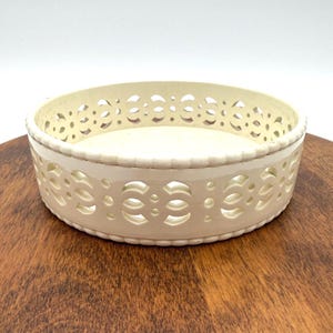 May include: Off-white, round decorative bowl with a pierced, geometric design around the sides. The bowl sits on a wooden surface. The bowl's interior is a solid off-white color.
