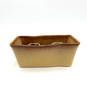 May include: A rectangular ceramic planter with a two-tone brown and tan glaze. The planter has a slightly irregular top edge and two circular openings. The exterior has a matte finish.