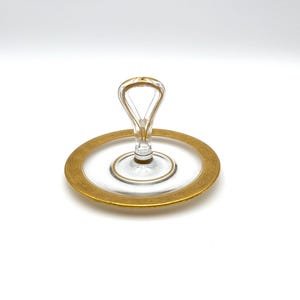 May include: A clear glass serving plate with a gold rim and handle. The plate is round, with a decorative gold border. The handle is arched and also trimmed with gold. The plate is designed for serving appetizers or desserts.