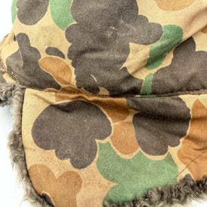 May include: A trapper hat featuring a camouflage pattern in shades of brown, tan, and green. The exterior fabric has a matte finish, while the interior and ear flaps are lined with a thick, textured gray faux fur material for insulation.