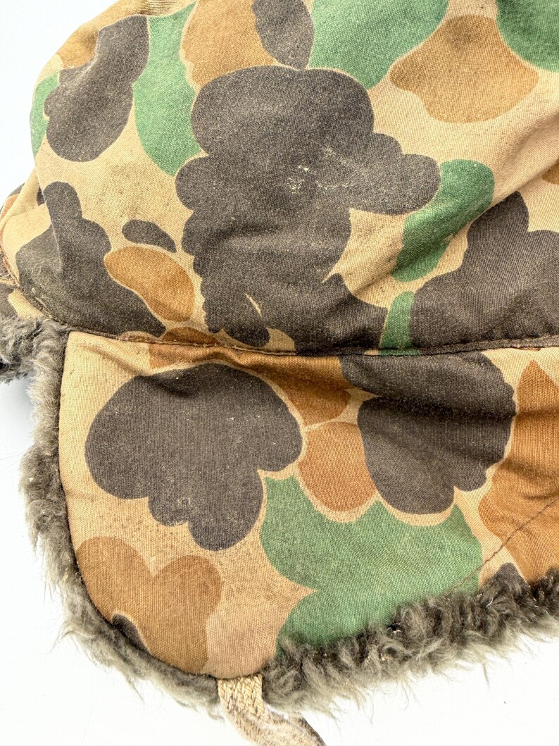 May include: A trapper hat featuring a camouflage pattern in shades of brown, tan, and green. The exterior fabric has a matte finish, while the interior and ear flaps are lined with a thick, textured gray faux fur material for insulation.