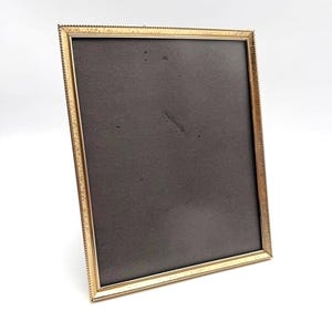 May include: A rectangular picture frame with a metallic gold finish and a textured, beaded edge detail. The frame contains a dark gray, solid-colored backing board that is visible through the center opening of the frame structure.