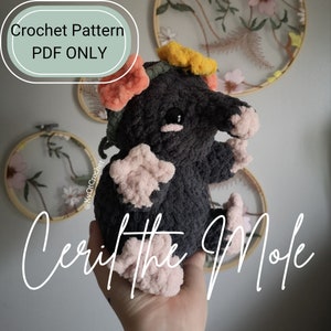 May include: Crochet pattern PDF for a cute gray mole amigurumi toy with pink and orange flower accents. The toy is being held in a hand. The text "Cerit the Mole" is written below the toy.