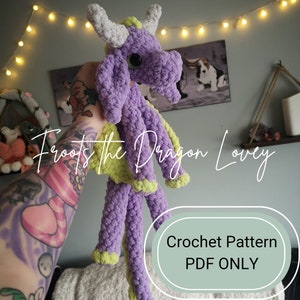 Op de afbeelding: Haakpatroon voor een paarse en groene draken knuffel. Het patroon is alleen beschikbaar als PDF-download. De afbeelding toont een gehaakte draken knuffel die door een persoon wordt vastgehouden.