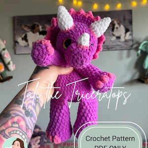 May include: A handmade, crocheted triceratops dinosaur in shades of purple and pink. The plush toy has white horns, a frill, and small, round eyes. The text "Tali the Triceratops" is written on the image, along with "Crochet Pattern PDF ONLY".