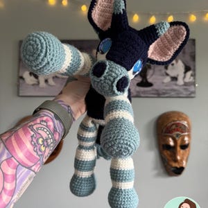 May include: A handmade crochet plush toy featuring a dark navy blue body, head, and ears with light pink inner ear details. The limbs are striped with light blue and white yarn. The creature has large, glossy blue eyes and a dark navy blue snout.