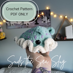 May include: Crochet pattern for a sea slug amigurumi toy named "Suds the Sea Slug". The toy is light blue with purple spots and white frills. The pattern is available as a PDF download only.