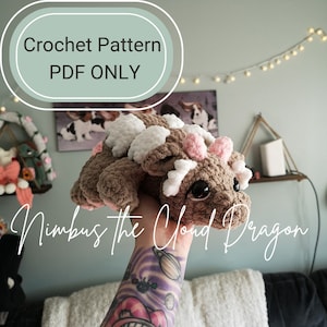 Może przedstawiać: Wzór na szydełku PDF dla amigurumiego w kształcie brązowego i białego chmurnego smoka. Smok ma różowe akcenty i duże czarne oczy. Tekst "Nimbus the Cloud Dragon" jest napisany pod smokiem.