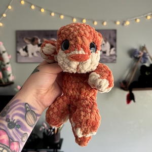 Puede incluir: Un juguete de peluche de color naranja y blanco tejido a crochet con forma de cachorro de tigre. El juguete tiene ojos grandes y negros y un hocico blanco.