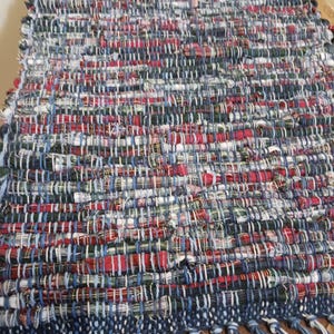 May include: A rectangular, handwoven rug with a mix of red, blue, white, and green threads. The rug has a fringed edge in shades of blue and white. The woven pattern creates a textured surface, suitable for a hallway or entryway.