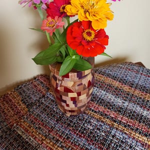 May include: A wooden vase constructed from small, multi-colored blocks of wood in various shades of brown, tan, and purple. The vase holds a vibrant bouquet of fresh zinnia flowers in shades of bright red, orange, yellow, pink, and pale green.