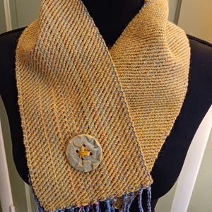 May include: A handwoven scarf in a golden-yellow hue with a textured, diagonal weave. The scarf features a large, decorative button and fringed ends with blue and purple threads. The scarf is displayed on a black mannequin.